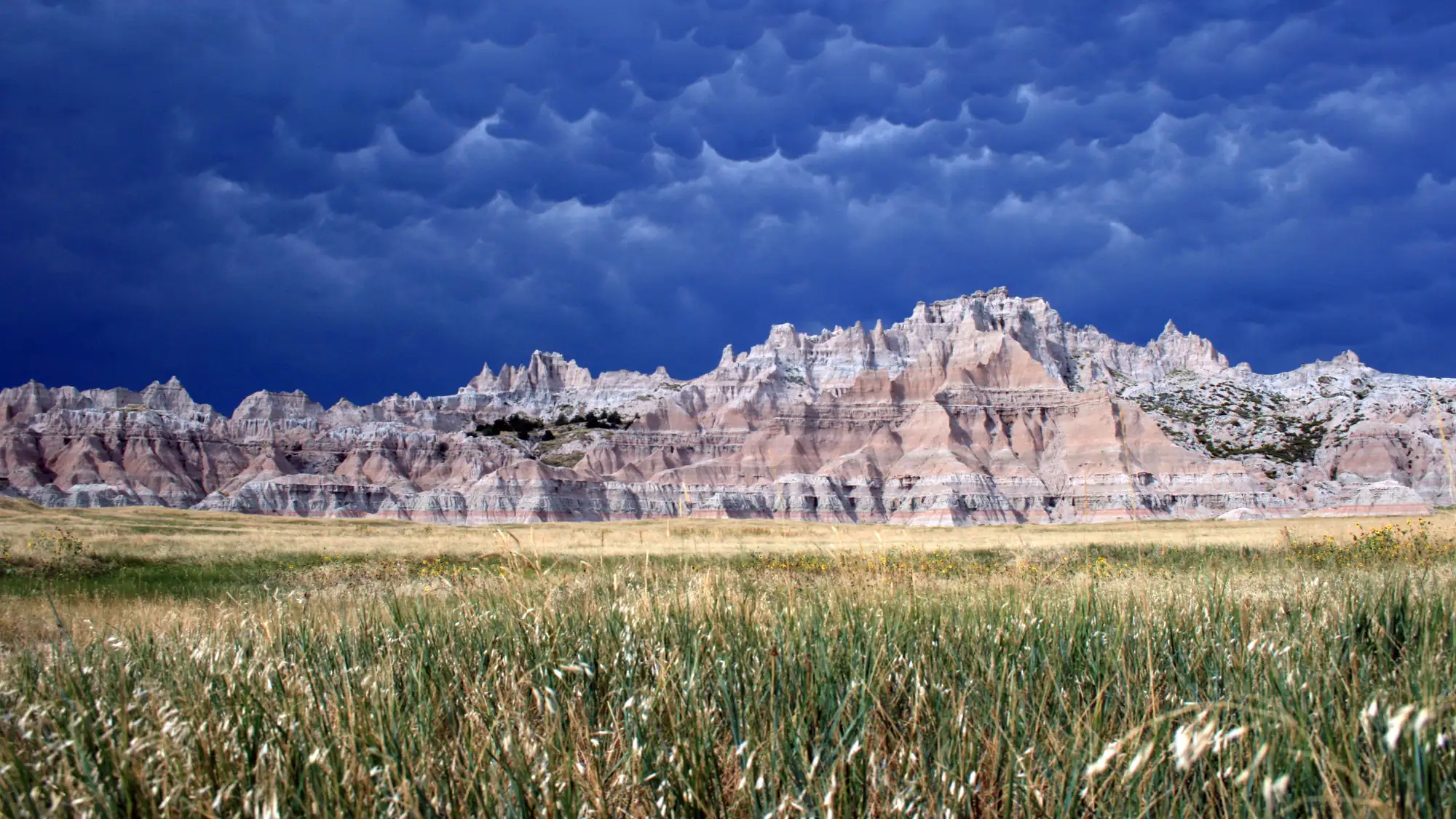 Layered badlands formations behind fields of green grass under cloudy and billowing clouds.
