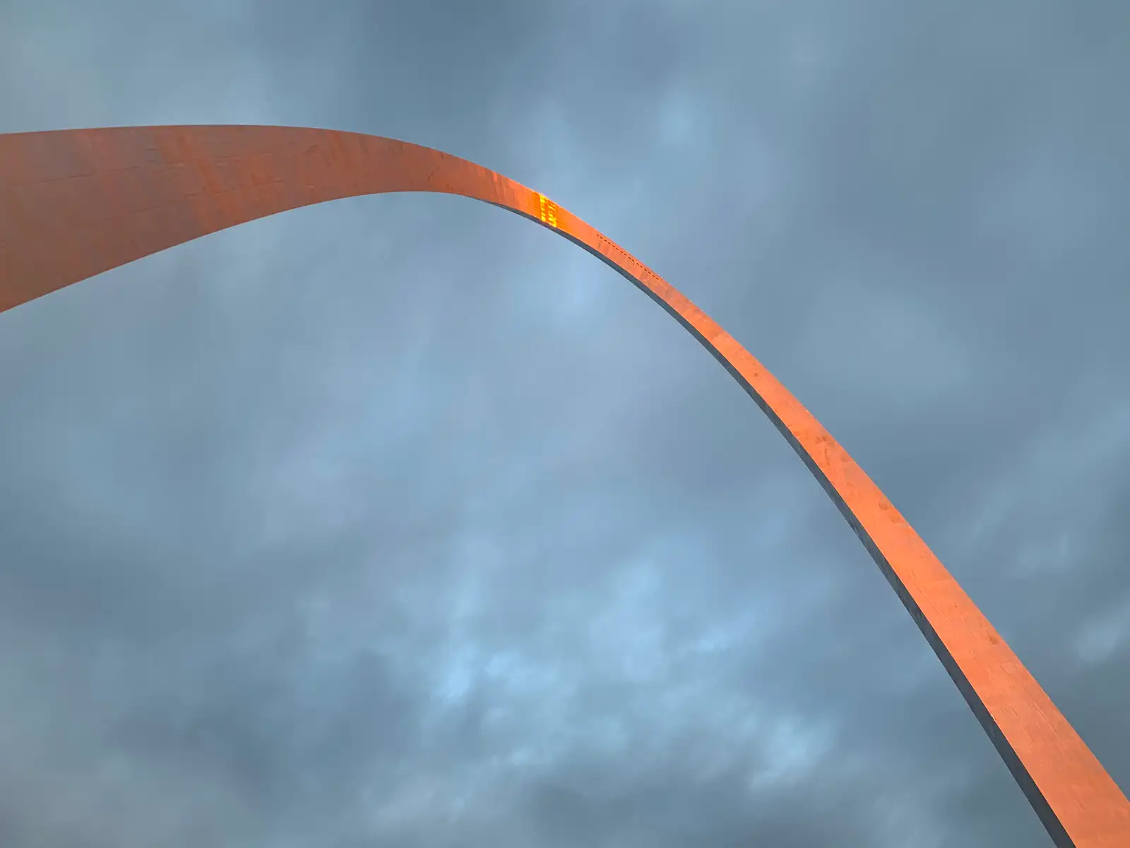looking up at the Gateway Arch against a cloudy blue sky with a orange reflection at the top