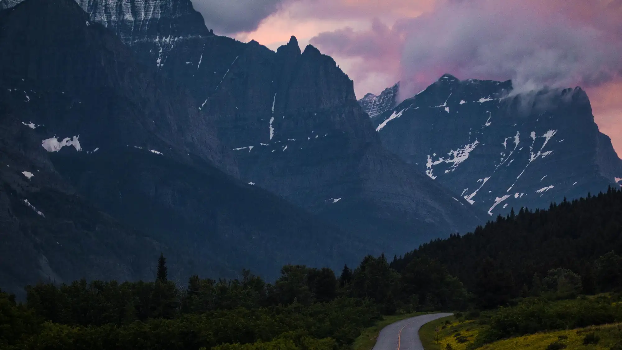 Jagged peaks rise out of a forested valley and an empty road curves off into the distance.