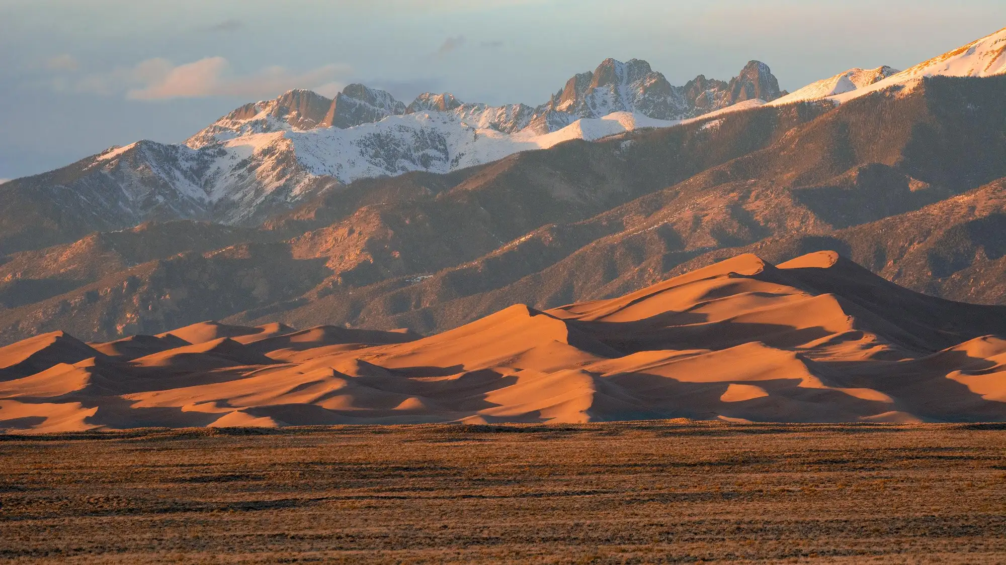 Grasslands, large dunes, and snow-capped peaks at sunset