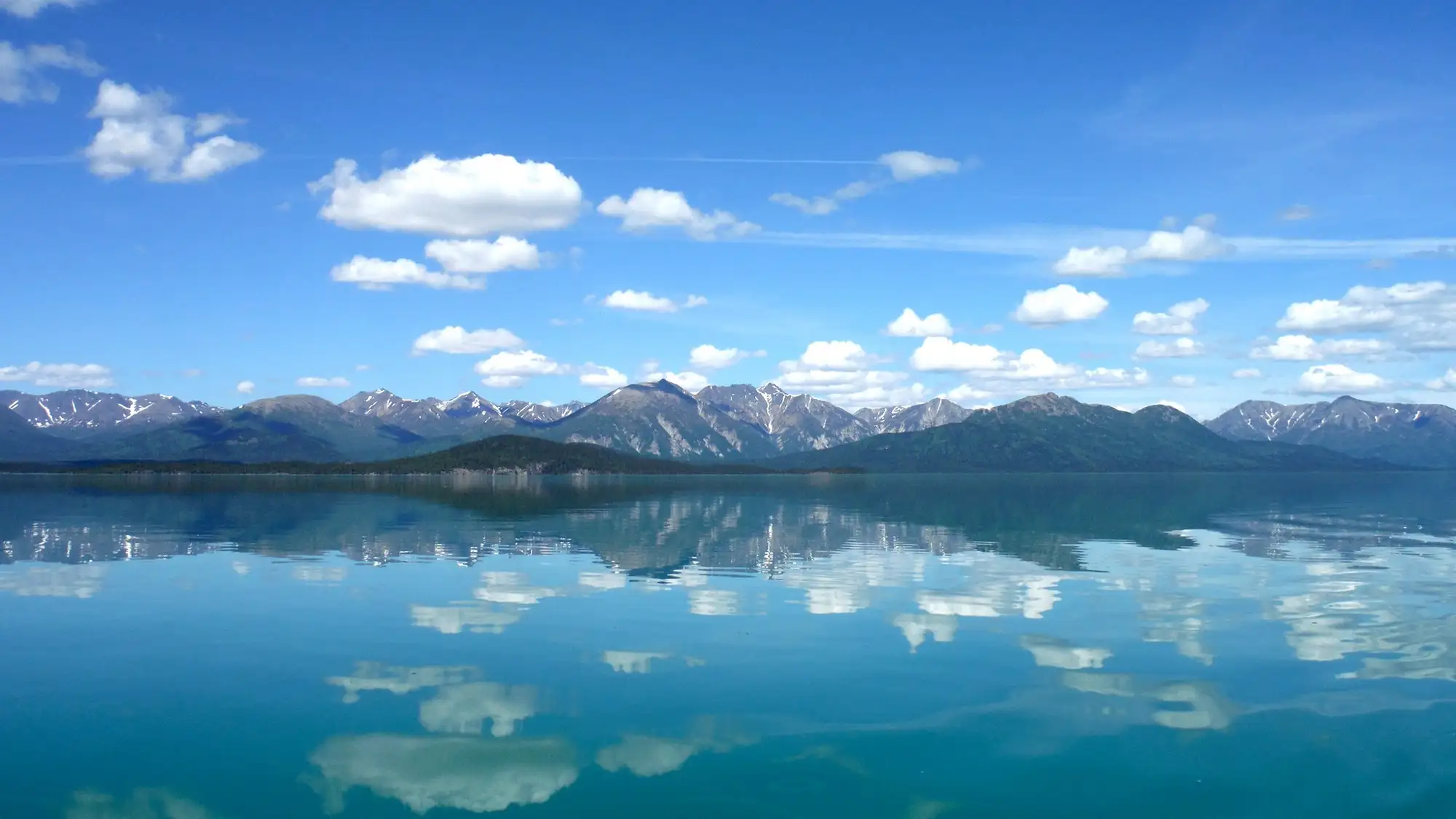 Photo of blue sky with fluffy white clouds reflect in calm lake with mountains in the background.
