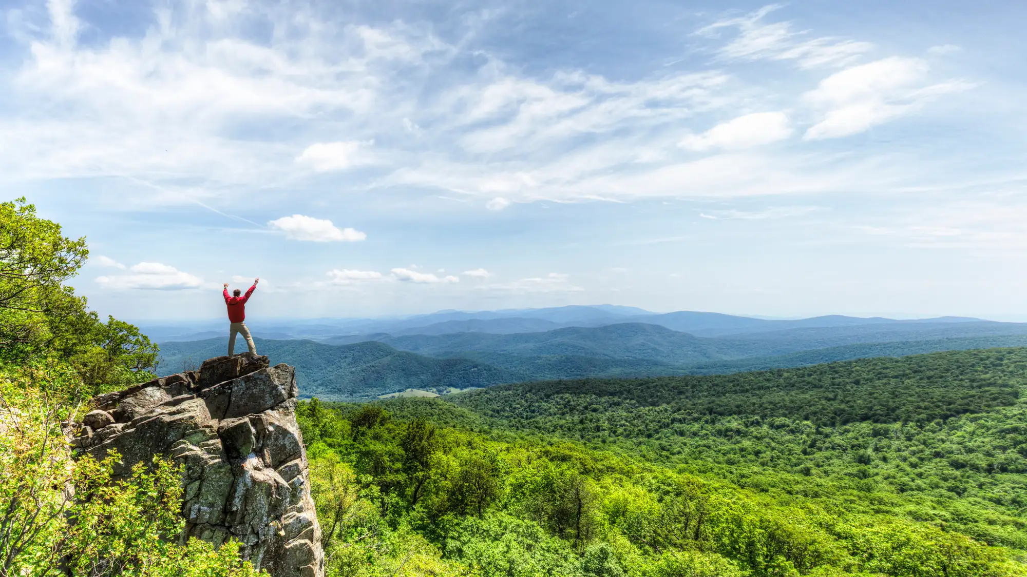A man stands on a rocky outcrop overlooking the receding mountains.