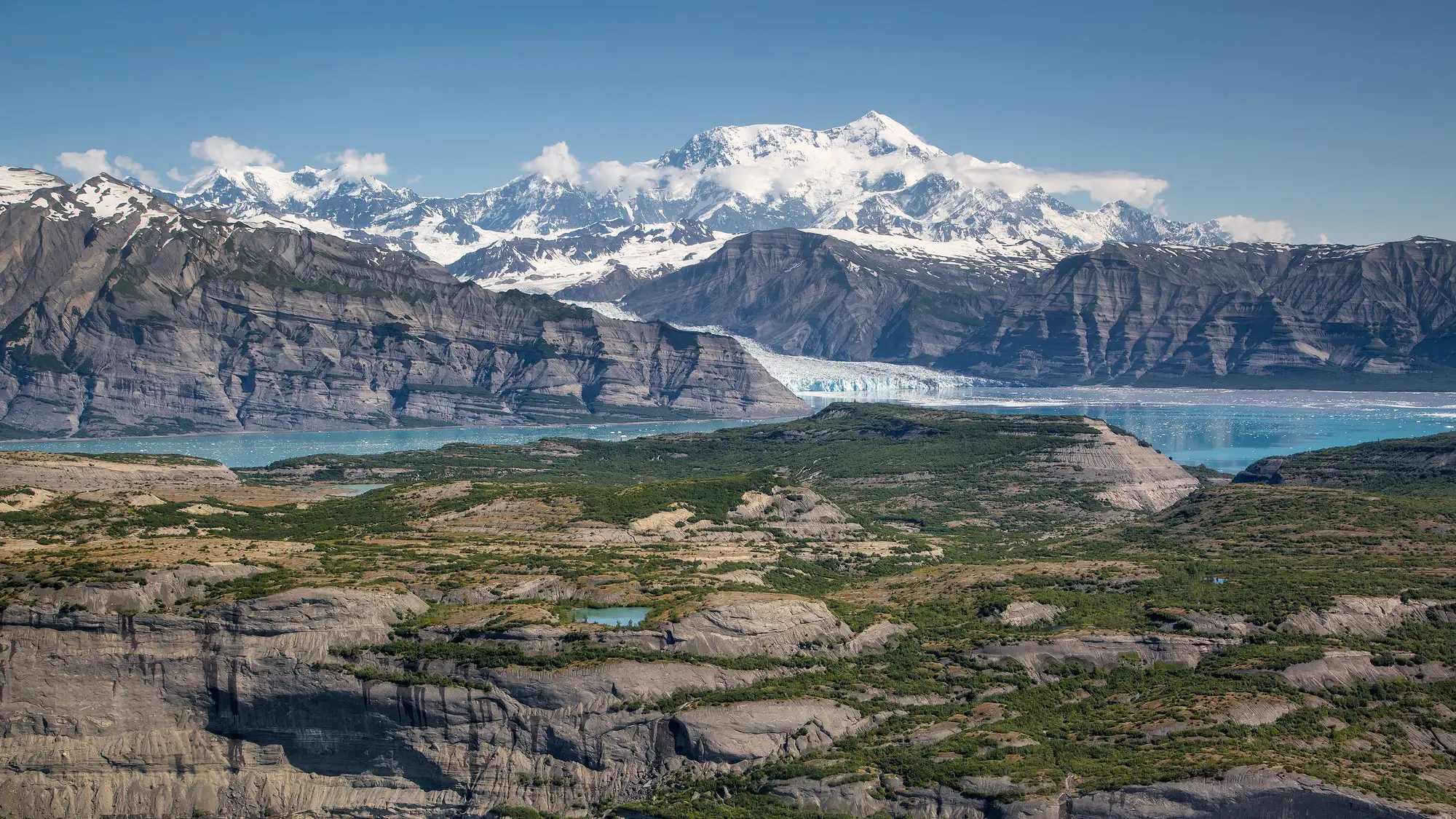 Glaciers loom over the ocean with large snowy mountains rising into blue skies